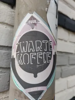 A close-up shot of a Zwarte Koffie sticker. The sticker is round with a black background and white text. The text says "Zwarte Koffie" which is Dutch for black coffee.  The sticker also has the words "Puur" and "Club Up" on it. The sticker appears to be on a pole outdoors.