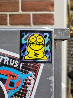 A yellow cartoon character with stitched mouth and closed eyes, smiling happily. The sticker has a colorful, geometric background. The sticker is placed on a grey metal box.