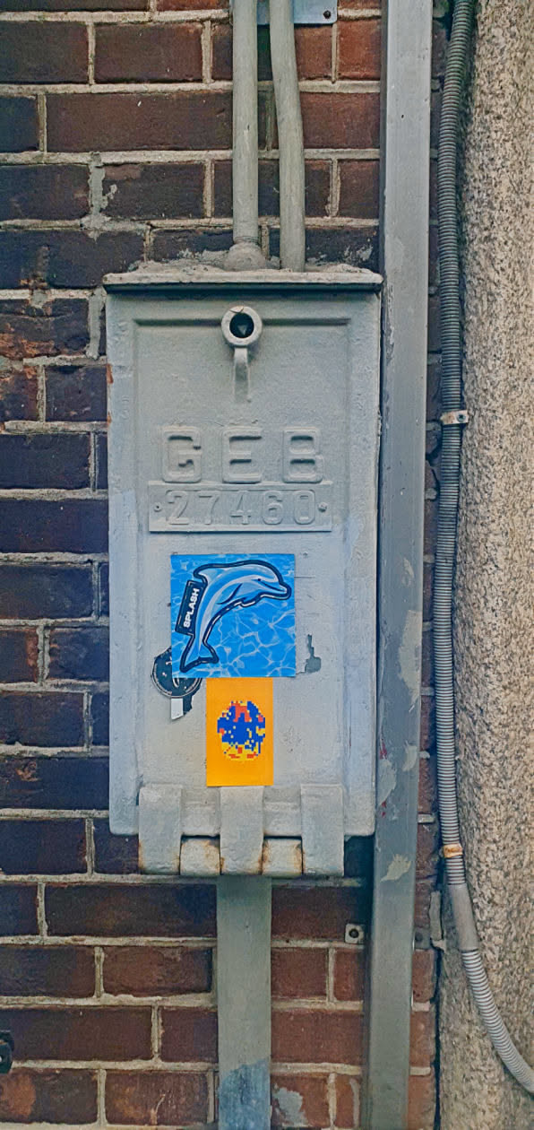 A dolphin sticker is placed on a metal electrical box. The sticker features a drawing of a dolphin over light blue water with the text 'SPLASH' on the left side. Below this sticker is a small sticker with a mosaic image of blue, red and yellow on an orange background. The electrical box is old and gray with the letters 'GEB' and the numbers '27460' embossed on the front.