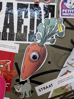 Street sticker A sticker of a carrot with one googly eye. The carrot is orange with green leaves on top. There is a small yellow sticker on the leaves with the text '@Boodzz'.