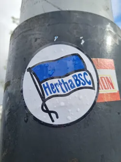 A round sticker featuring a stylized Hertha BSC flag. The flag is blue and white with the text "Hertha BSC" written on it. The sticker is slightly wet, with several water droplets visible on its surface.