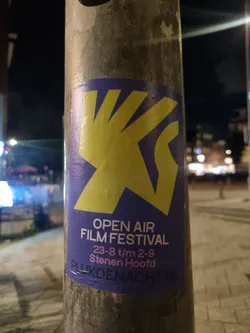 A purple and yellow sticker advertising an open air film festival in Stenen Hoof from August 23rd to September 9th. The sticker features a stylized hand graphic and the website Plukdenacht.nl.
