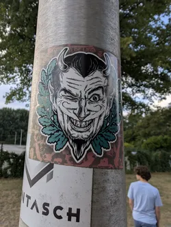 Street sticker A rectangular sticker depicting a black-and-white winking devil with horns and a goatee, encircled by a teal oak leaf wreath on a distressed red background.