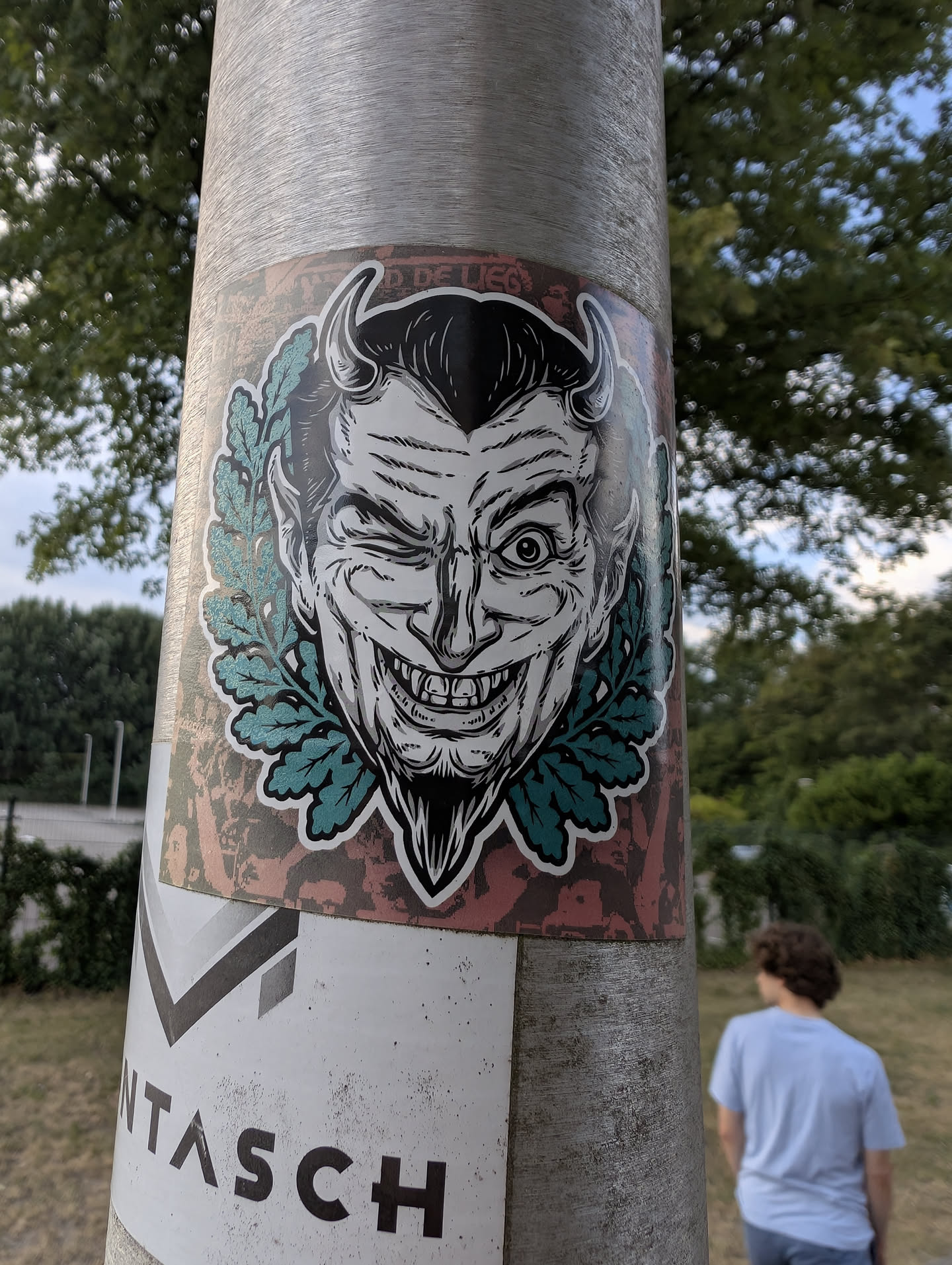 A rectangular sticker depicting a black-and-white winking devil with horns and a goatee, encircled by a teal oak leaf wreath on a distressed red background.