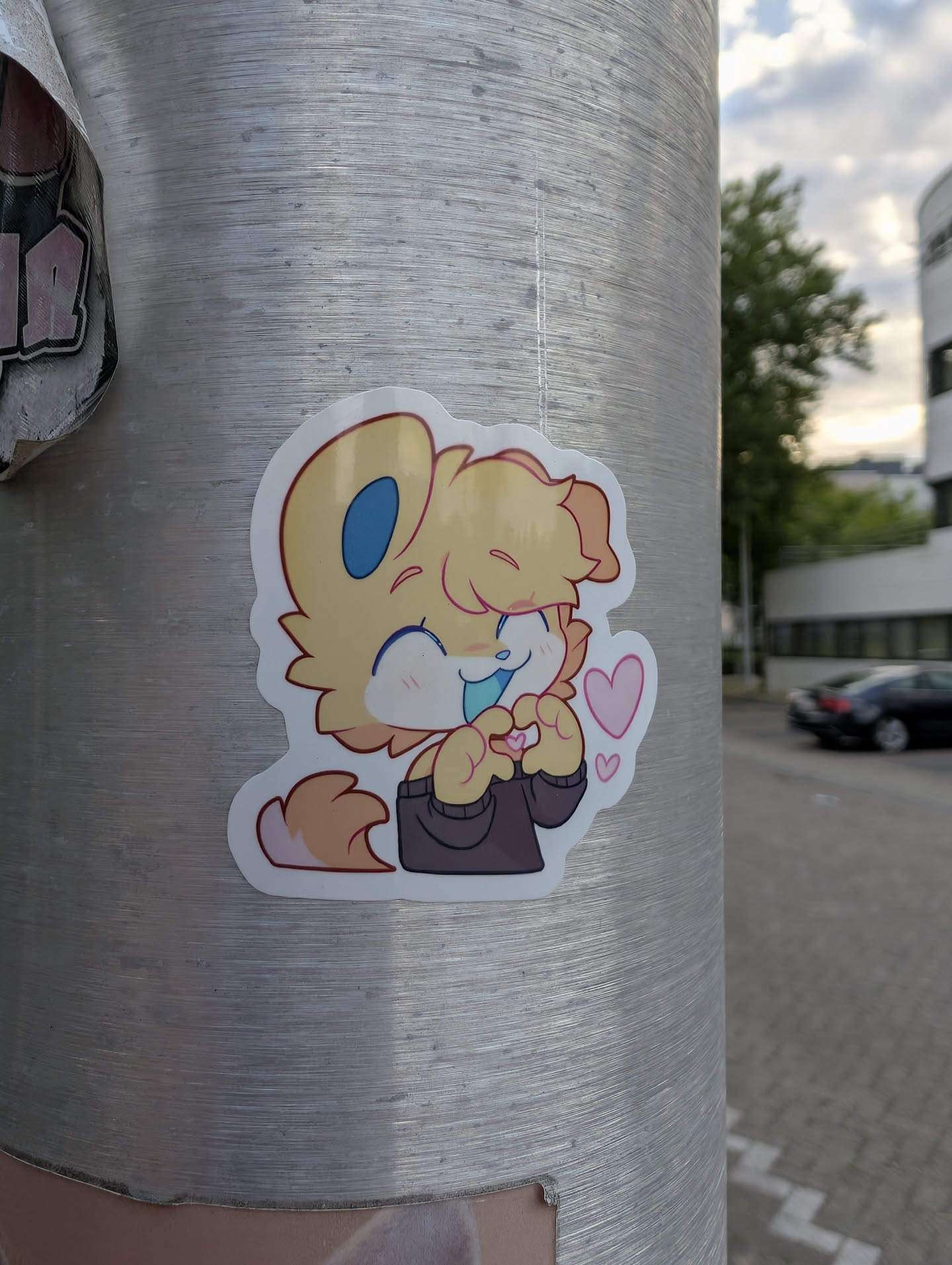 A die-cut sticker of a chibi-style yellow furry character with blue inner ears and tongue. The character wears a dark sweater, smiles with closed eyes, and forms a heart shape with its paws, flanked by small pink hearts.