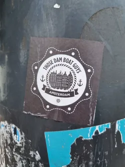 A round sticker with a dark background featuring the text "Those Dam Boat Guys" in a circular banner above an illustration of Amsterdam buildings. Below the illustration, the word "Amsterdam" is written. The sticker is adorned with anchors and nautical rope designs, giving it a nautical theme. The sticker is slightly damaged and shows signs of wear.