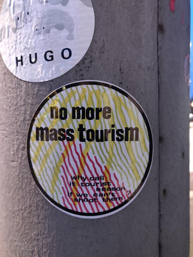 Street sticker Round sticker with a black outline and a colorful striped pattern. It displays the text "no more mass tourism" in bold black letters. Below it, smaller text reads: "why call it tourist season if we can't shoot them".