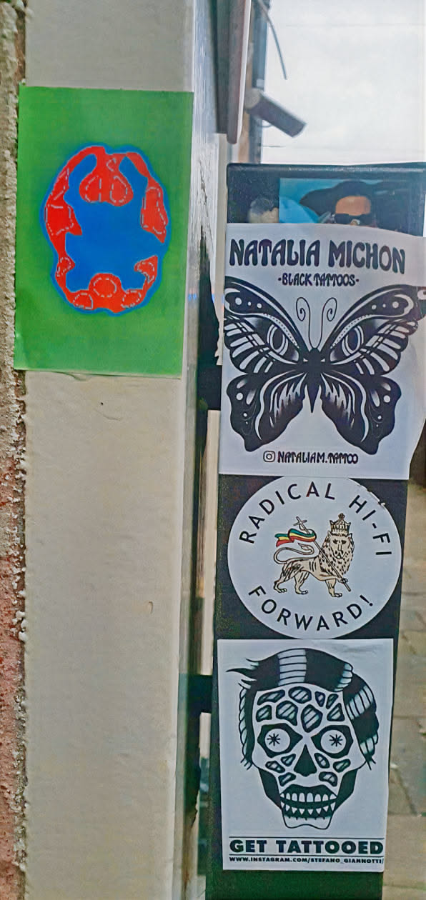A close-up of a pillar adorned with stickers and posters. The second sticker promotes 'NATALIA MICHON -BLACK TAMOOS-', it features a butterfly graphic and the Instagram handle @NATALIAM.TAMOO. Next, there's a sticker with a lion and the text 'RADICAL HI-FI FORWARD!'. Finally, there is a skull sticker with the text 'GET TATTOOED' and the Instagram handle 'WWW.INSTAGRAM.COM/STEFANO GIANNOTTI'. There's also an abstract drawing on a green background above the Natalia Michon sticker.