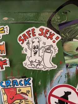 Street sticker The sticker features a cartoon character, resembling a phallus with arms and legs, in a simplified, bold black outline style. The character has wide eyes, a smiling mouth, and is pointing its index finger upwards with a playful expression. It is holding a condom package in its other hand. The text 'SAFE SEX!' is prominently displayed above the character in bold, red, block letters with a black outline. The overall impression is energetic and message-driven, consistent with a pop art or graffiti-inspired aesthetic.