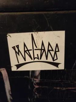 Street sticker A white square sticker with black lettering. The word 'MALARA' is written in a stylized, almost graffiti-like font. The letters are sharp and angular, with a crown-like shape above them.  The sticker appears to be adhered to a dark surface.