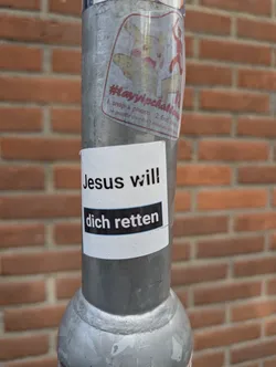 A white sticker with black text affixed to a metal pole. The text reads "Jesus will dich retten". The phrase translates to "Jesus will save you". There is another sticker above it. It appears to be a Pikachu character.