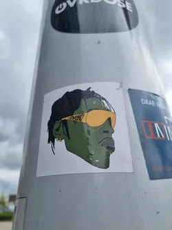 Street sticker This sticker shows a stylized illustration of a person's head and shoulders, possibly a celebrity or fictional character.  The individual has short dark hair, gold-framed yellow sunglasses with what appears to be a small inscription on the frame, and is depicted in shades of green and black. The style is a bit cartoonish, almost like a vector graphic.