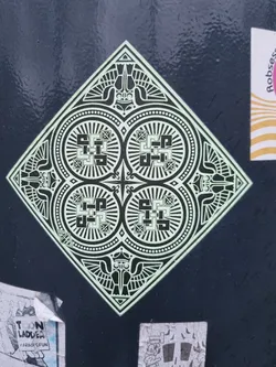 Street sticker A diamond-shaped sticker featuring a symmetrical design with four circular motifs and winged creatures at each corner. The pattern is primarily black and white, with a light green or beige background. The style is reminiscent of ancient or tribal art.