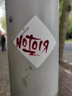 Street sticker A diamond-shaped sticker with the text \"NoTo19\" written in maroon paint. The sticker is affixed to a gray pole.  A small EKOPLAZA logo is visible in the upper part of the sticker.