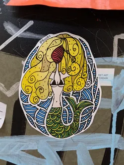 Street sticker A sticker featuring a stylized mermaid with yellow hair, a red top, and a green scale tail.  The design is intricate and colorful, with blue and yellow waves surrounding the mermaid. The artist's Instagram handle is visible in the upper right corner of the sticker.