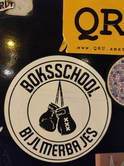 Street sticker Round sticker with a black and white illustration of a pair of boxing gloves hanging from a rope. The text "Bokschool Bijlmerbajes" is written around the image of the gloves. 