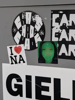 Street sticker A square sticker featuring a green-tinted close-up portrait of a woman. Bold orange text across her face reads 'A GIRL WITH A GUN', with a small red social media handle printed at the bottom.