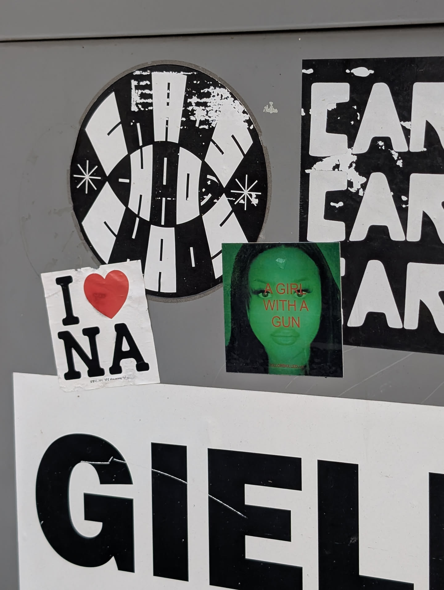 A square sticker featuring a green-tinted close-up portrait of a woman. Bold orange text across her face reads 'A GIRL WITH A GUN', with a small red social media handle printed at the bottom.