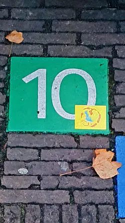 Street sticker The image shows a green square with a large white '10' painted on it, placed on a brick surface. Next to the number '0' is a small, yellow square sticker with a partially obscured image that vaguely resembles a world-like logo. A few dry leaves are scattered around the area.