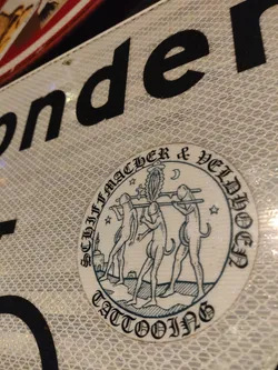 Street sticker A round sticker with a blue and white design depicting three figures carrying a pole with a plant on top. The text around the design says "Schiffmacher & Veldwesen Tattooing". The style is reminiscent of medieval or renaissance woodcuts.