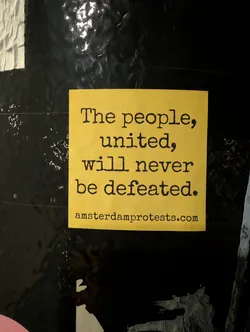 A square-shaped sticker with a bright yellow background and black text. The main message, 'The people, united, will never be defeated.', is prominently displayed in a distressed, typewriter-style serif font, centered on the sticker. Below this, in a smaller font of the same style, is the website 'amsterdamprotests.com'. The sticker is adhered to a dark, possibly metallic or painted, textured surface with some reflective highlights.