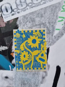 Street sticker A sticker featuring a floral design in yellow and blue, with hands reaching towards a central flower. The text 'Reach Out for Ukraine!' is at the bottom. The style is reminiscent of folk art or naive art.