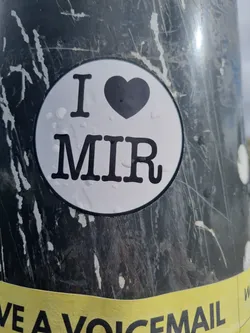 Street sticker A round sticker with the text "I ♥ MIR" in black on a white background. The sticker is affixed to a dark gray surface with scratches.