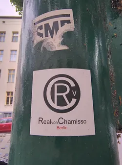 Street sticker The sticker features a black circular design with the letters 'R' and 'V' inside, above the text 'Real von Chamisso' in black, with 'Berlin' underneath in red. It is adhered to a green pole.
