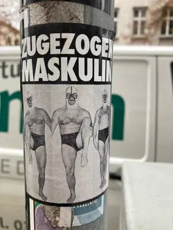 Street sticker The sticker prominently displays a stark black and white illustration of three muscular, bare-chested male figures. Each figure wears a distinctive full-face mask, resembling a wrestling or gimp mask, with circular cutouts for eyes and mouths. They are clad only in dark briefs. The central figure is depicted front-facing, while the two figures on either side are slightly angled, all striking a somewhat unusual pose with one leg lifted or bent. Above the figures, bold white sans-serif text on a solid black background reads 'ZUGEZOGENE MASKULIN'. The overall aesthetic is grainy, suggestive of a photocopied or roughly printed image, and is affixed to a textured, cylindrical pole with visible signs of wear and slight peeling.