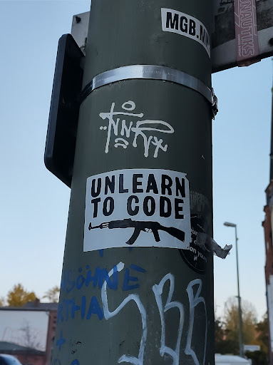 Street sticker A sticker with the text "Unlearn to Code" and an image of an AK-47 rifle. The sticker is affixed to a pole with other graffiti and stickers.