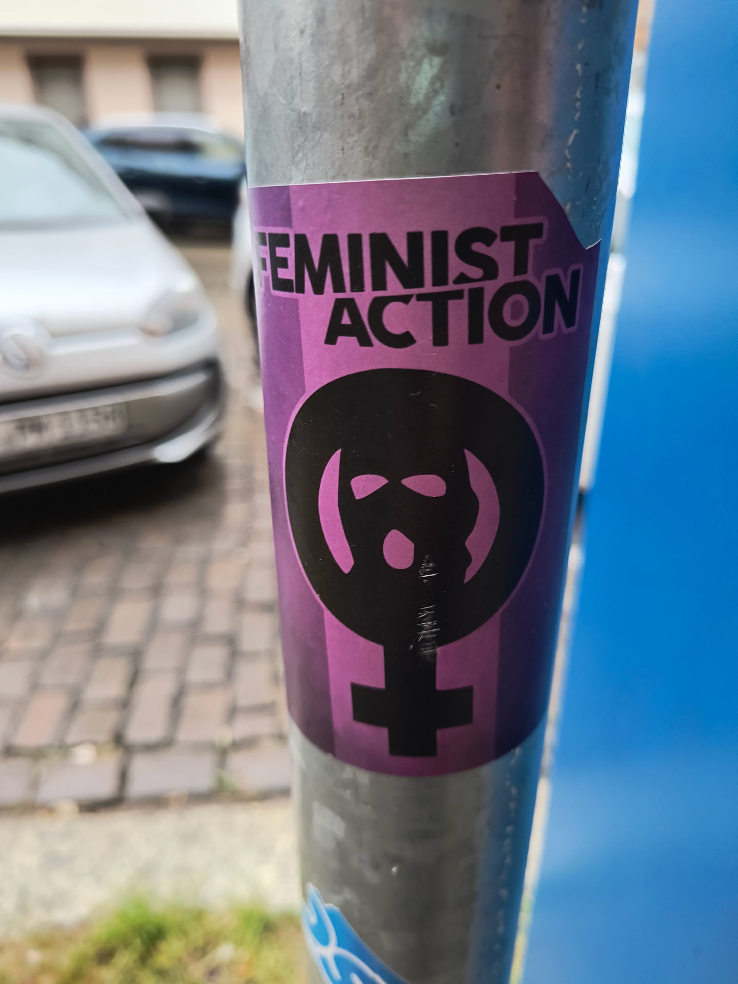 The sticker is predominantly purple with a gradient effect, featuring black text and a prominent black symbol. The text at the top reads 'FEMINIST ACTION' in bold, sans-serif capital letters. Below the text is the Venus symbol (♀), commonly associated with femininity and feminism, with a stylized, almost mask-like face superimposed within the circle of the symbol. The sticker appears to be applied to a metal pole.