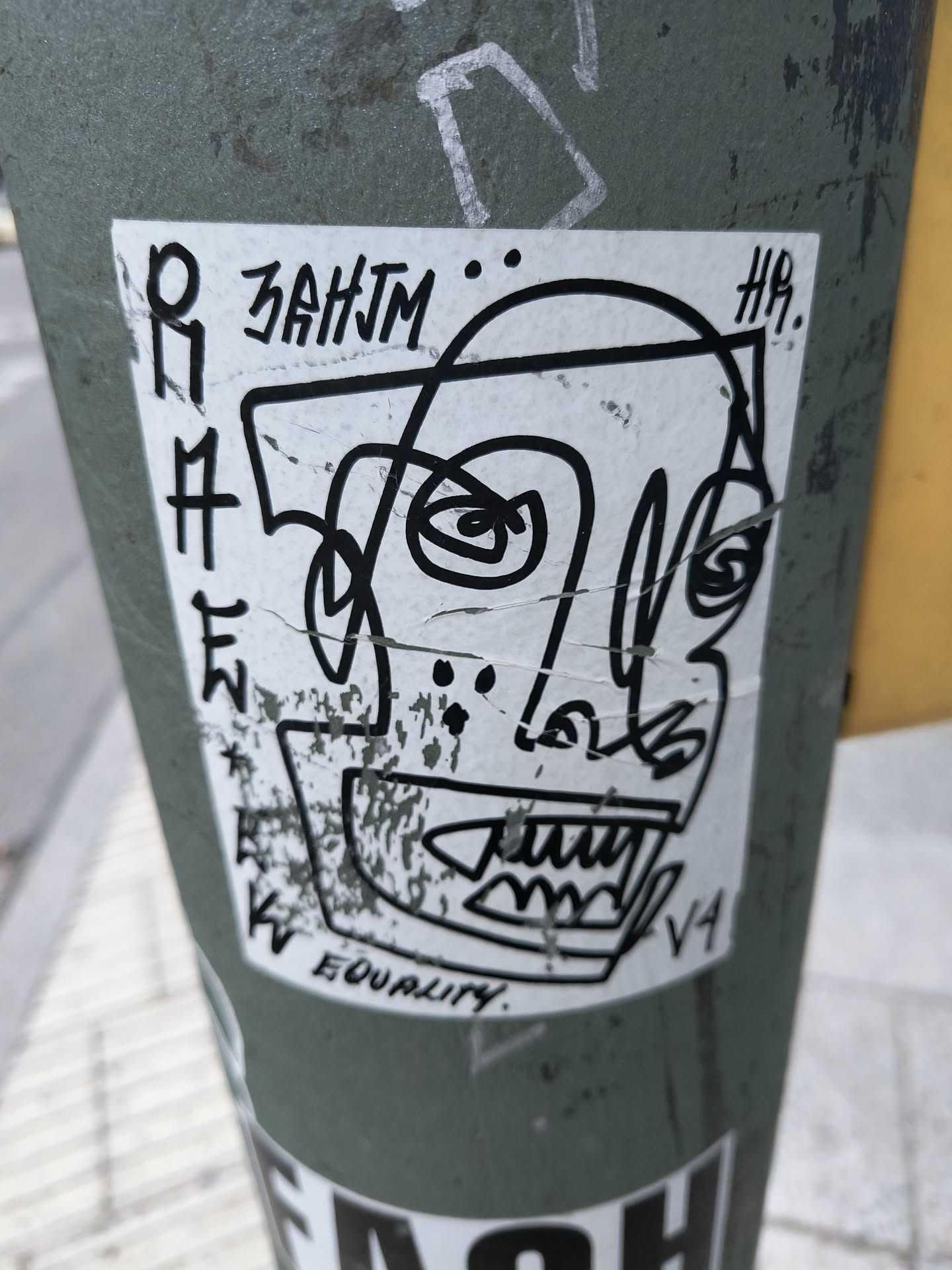 The sticker features a black and white, abstract depiction of a face with a distorted, angular head. The eyes are represented by swirling lines and dots, and the mouth is a jagged, toothy grin. The overall style is rough and expressive, with thick black lines. Text elements are present around the face, including what appears to be a signature or tag. The sticker itself shows some signs of wear and tear, with creases and minor damage.