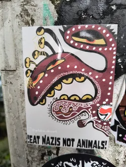 Street sticker This sticker depicts a cartoonish creature with a maroon/red body covered in white spots, along with yellow eyes and teeth. It has several tentacle-like appendages on its head. A smaller design below the creature resembles a stylized face. The phrase "EAT NAZIS NOT ANIMALS!" is prominently displayed at the bottom. There is also a red and white symbol resembling an anti-fascist action logo. The artist's Instagram handle is present as well.
