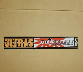 A horizontal sticker with the word "ULTRAS" in orange on a black background. Next to it is a red and white Japanese rising sun design, followed by the words "ÜBERKLEBT" in red with a crown on top, and "SUPPORT FRANKFURT" in smaller letters underneath. The sticker has a slightly worn look.