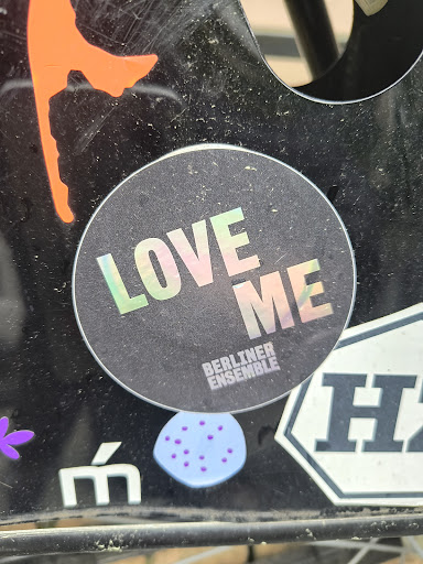The sticker features a black background with the text 'LOVE ME' in a holographic, rainbow-reflective font. Below this, the words 'BERLINER ENSEMBLE' are printed in smaller font. The sticker is adhered to a black surface.