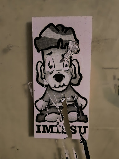 Street sticker A sticker featuring a cartoon character with a dog-like face, wearing a hat and sitting down. The character is drawn in black and white with a somewhat distressed style, and the sticker itself shows signs of wear and tear. The text "IMISU" is printed below the character.