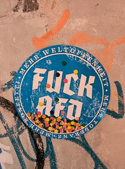 Street sticker The sticker is circular and predominantly blue with white text and accents. The text 'FUCK AFO' is prominently displayed in large, bold white sans-serif font, with the letters 'FUCK' on the top line and 'AFO' on the bottom. Scattered around the text and within the bottom portion of the sticker are numerous small, colorful circles resembling confetti, in shades of pink, yellow, orange, green, and blue. The outer edge of the sticker features text in white, which appears to be German: 'MEHR WELTOFFENHEIT • MEHR TOLERANZ • MEHR KONEETTI'. The blue background has a distressed, textured appearance, with white speckles and scratches, suggesting wear and tear. The sticker is affixed to a wall with visible graffiti in the background.