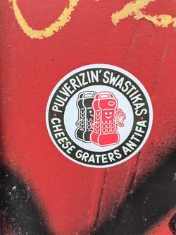 Street sticker A circular sticker featuring a bold, graphic design. The central image depicts two stylized, anthropomorphic cheese graters against a white background; one is black and the other, in front, is red with a friendly smiling face. The red grater appears to be actively 'pulverizing' a small, distorted red swastika symbol, indicating its destruction. Encircling the central image, along a thick black border, is white uppercase text. The overall aesthetic is clean and cartoon-like, emphasizing a strong political message.