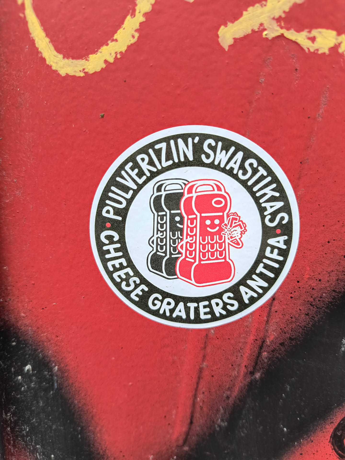 This is a round sticker on a red surface. The sticker has a white background with a black border. The text around the edge of the sticker reads "Pulverizin' Swastikas Cheese Graters Antifa". The image in the center of the sticker depicts three cheese graters. The cheese graters are in black, gray and red. The red cheese grater has a face on it and is holding a broken swastika.