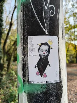 Street sticker A sticker featuring an illustration of a toddler wearing a crown and a dark collared garment. The style is dark and slightly whimsical, with small pink rose-like details on the garment.  Text on the sticker includes an unclear phrase and social media handles.