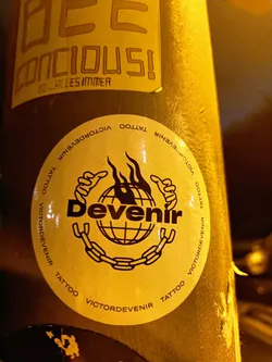 Street sticker Round sticker with the word 'Devenir' in bold text inside a circular emblem featuring a globe encircled by a chain, all set against a yellow background. The text 'VictorDevenir Tattoo' is repeated around the outer edge of the circle.  The style is bold and graphic.