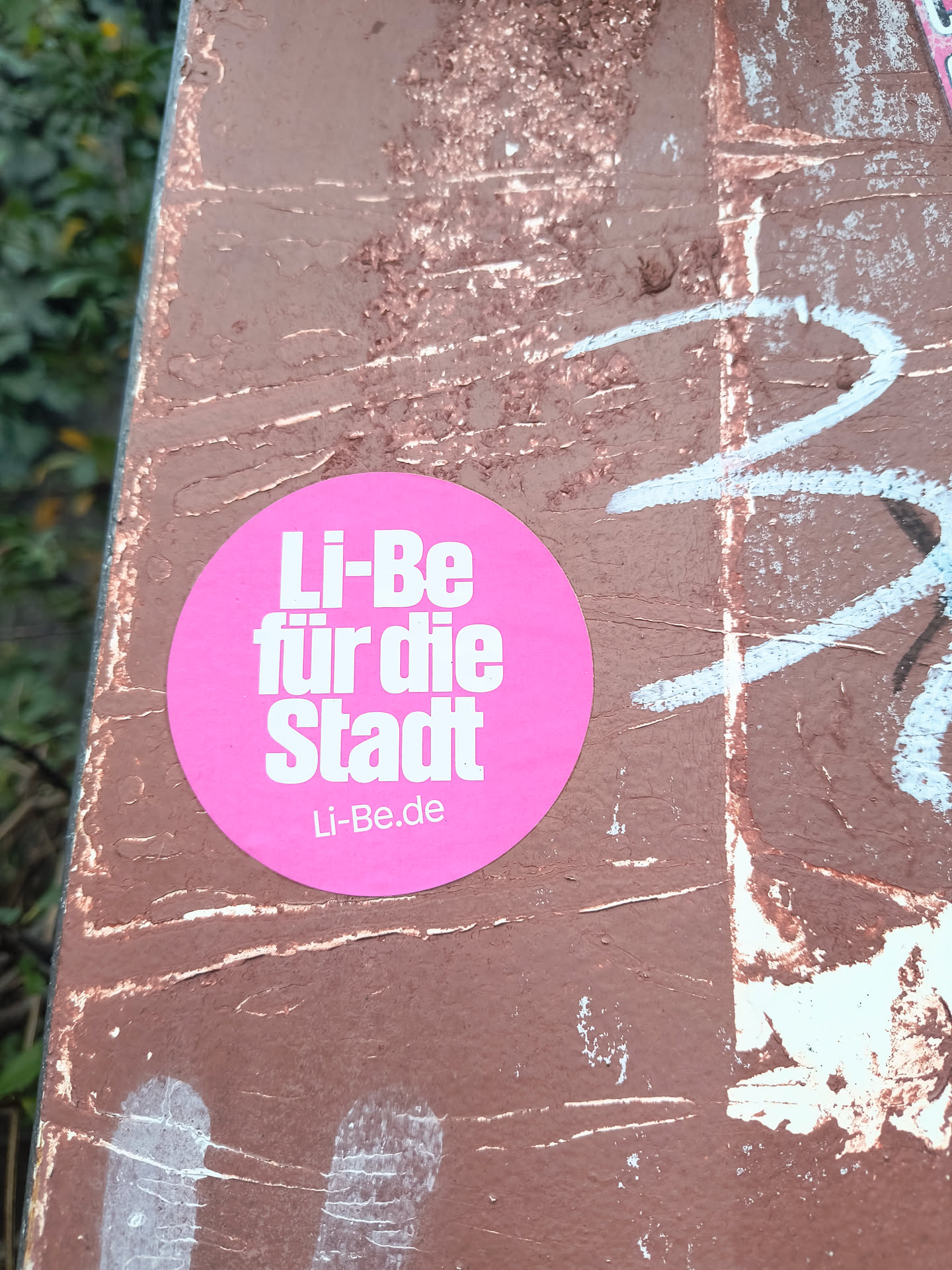 A bright pink circular sticker is centrally placed on a weathered, brown surface that has white and blue graffiti marks. The sticker features bold, white sans-serif text. The top line reads 'Li-Be', the second line reads 'für die', and the third line reads 'Stadt'. Below this, in a smaller font, is 'Li-Be.de'. The sticker appears to be in good condition, with clean edges and clear text.