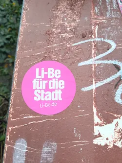Street sticker A bright pink circular sticker is centrally placed on a weathered, brown surface that has white and blue graffiti marks. The sticker features bold, white sans-serif text. The top line reads 'Li-Be', the second line reads 'für die', and the third line reads 'Stadt'. Below this, in a smaller font, is 'Li-Be.de'. The sticker appears to be in good condition, with clean edges and clear text.