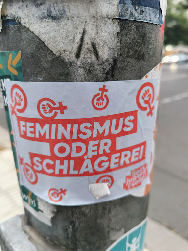 Street sticker A sticker expressing a choice between feminism and violence. It features the text "FEMINISMUS ODER SCHLÄGEREI" (Feminism or Violence) in red on a white background, along with several feminist symbols. The sticker is affixed to a pole, surrounded by other stickers and debris.