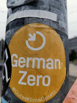 Street sticker Round yellow sticker promoting German Zero initiative.  It features a circular arrow symbol and the text "German Zero" in white. A smaller text at the bottom reads  "#GutesKlimaGesetz".  The top of the sticker contains small text in German indicating that the sticker is vegan and good for everyone.