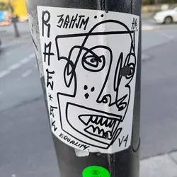 Black and white sticker featuring a stylized, abstract face with sharp lines and bold features. The style is reminiscent of cubism or graffiti art.  The sticker includes the text "ZAHJM" at the top and "EQUALITY" at the bottom. There are also some illegible markings next to the face illustration. The sticker is affixed to a pole.