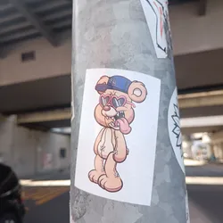 Sticker of a cartoon bear wearing heart-shaped sunglasses and a baseball cap. The bear has its tongue sticking out. 