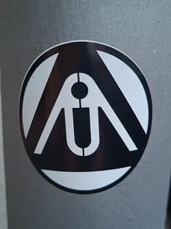 A round sticker with a black and white design. The logo appears to be an abstract symbol or emblem. The sticker is affixed to a gray surface, possibly a pole or a wall.