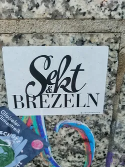 A sticker with the text "Sekt & Brezel" in a stylized font. The sticker is white with black text, and there is a small social media handle in the bottom right corner.