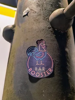 Street sticker A sticker featuring a stylized neon pink and blue rooster illustration inside a circle with the text ".BAR. ROOSTER."  The sticker is adhered to a dark gray, textured surface.